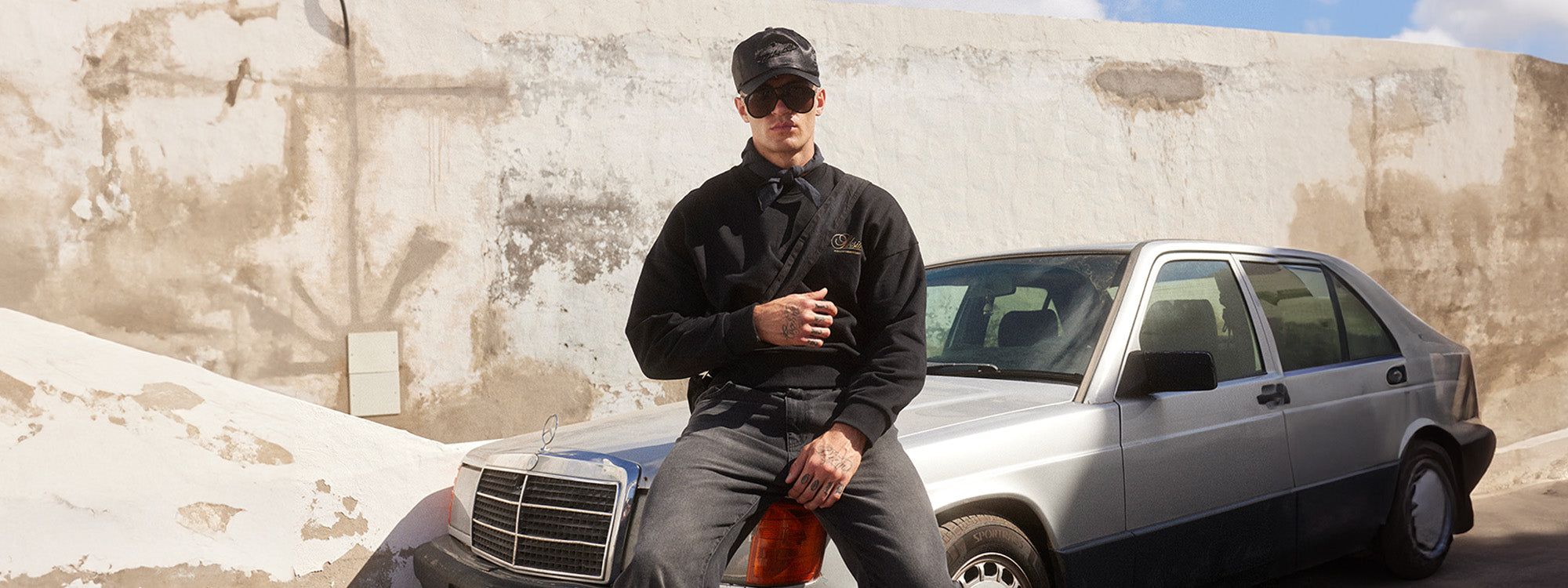 SikSilk New Arrivals. SikSilk model posing, sat on a car bonnet, wearing SikSilk Black Graphic Sweater, SikSilk Black Slim Fit Jeans & SikSilk Black Trucker Cap