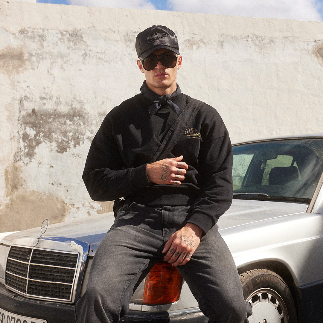 SikSilk New Arrivals. SikSilk model posing, sat on a car bonnet, wearing SikSilk Black Graphic Sweater, SikSilk Black Slim Fit Jeans & SikSilk Black Trucker Cap